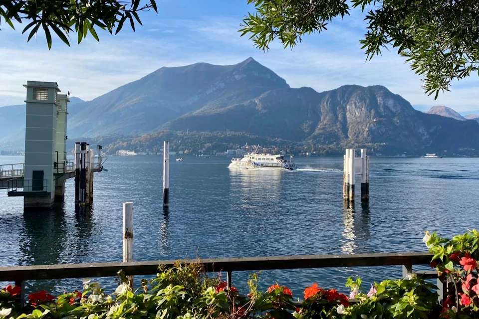 A boat travels across a lake surrounded by mountains; colorful flowers and greenery frame the foreground.