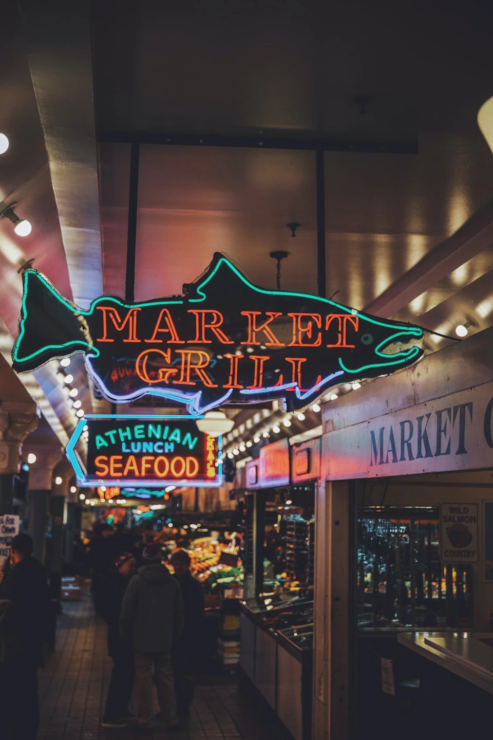 A market hallway with neon signs for 'Market Grill' and 'Athenian Lunch Seafood,' under a ceiling with string lights.