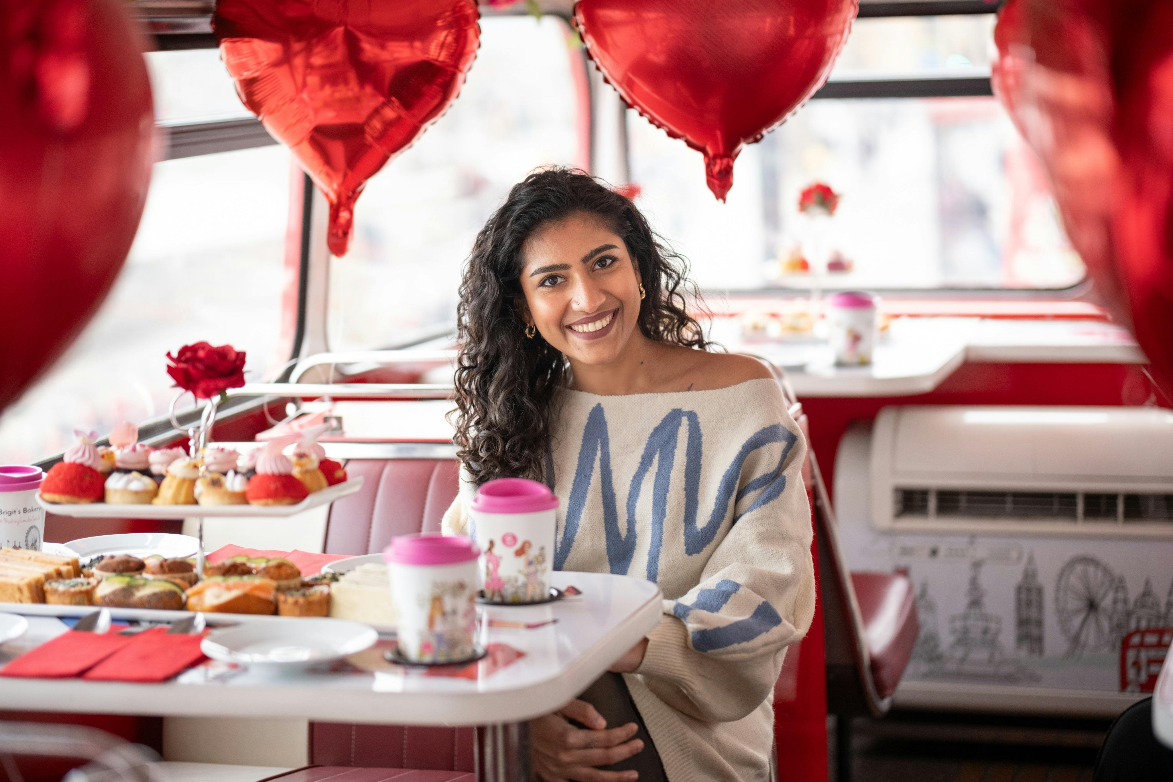 En kvinde sidder ved et bord i en hyggelig spisestue med rød indretning og smiler. Bordet har plads til mad, drikkevarer og hjerteformede balloner.