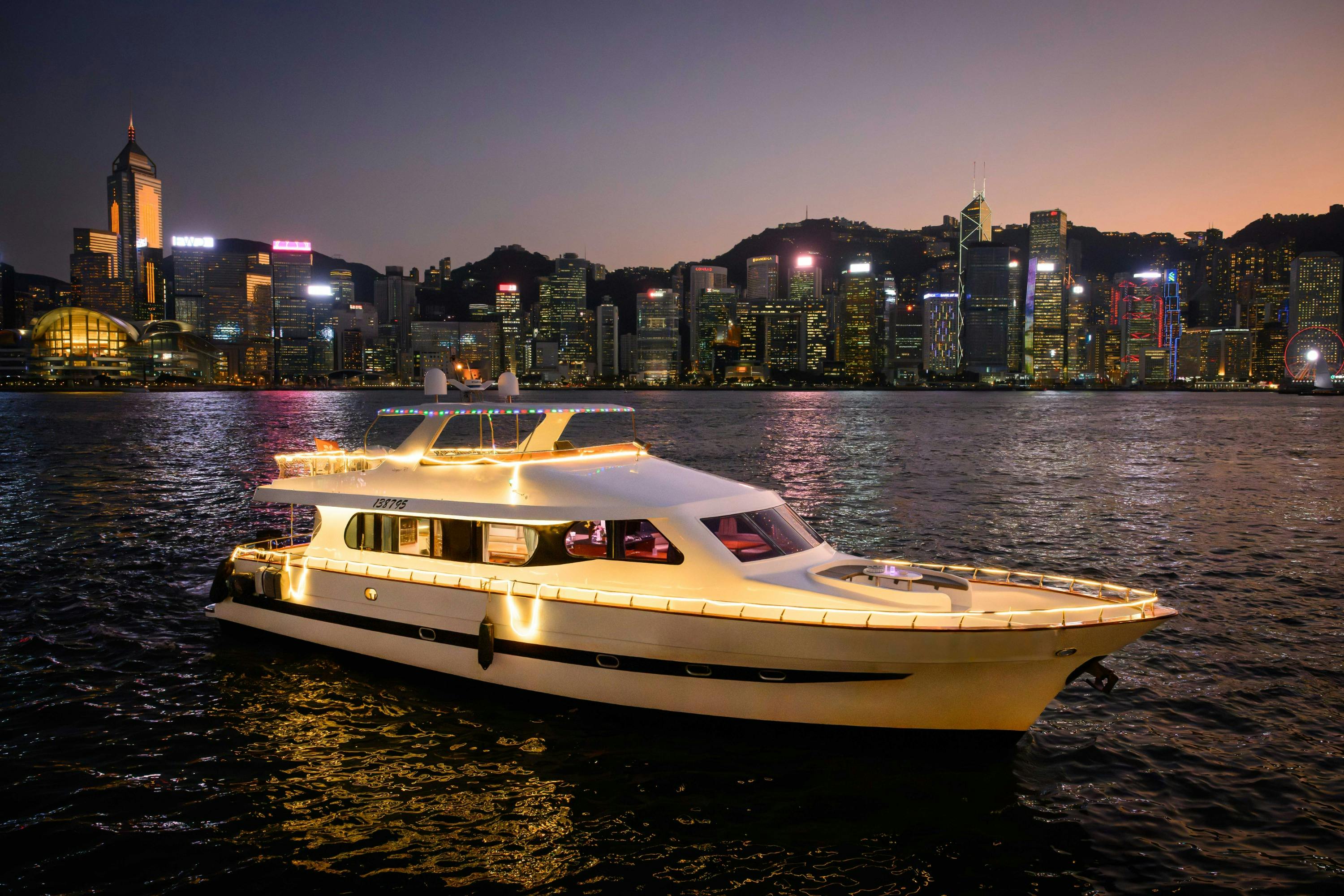 A lit-up yacht on water at night with a city skyline and illuminated buildings in the background.
