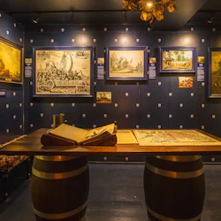 En museiutställning med historiska målningar på mörka väggar, ett centralt träbord med en öppen bok och en karta.