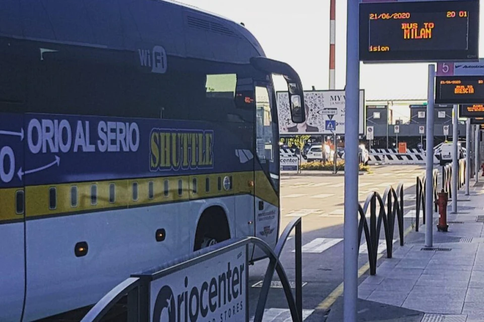 A shuttle bus parked at a station with a digital sign displaying the date and time. Nearby is a sign for "Orocenter."
