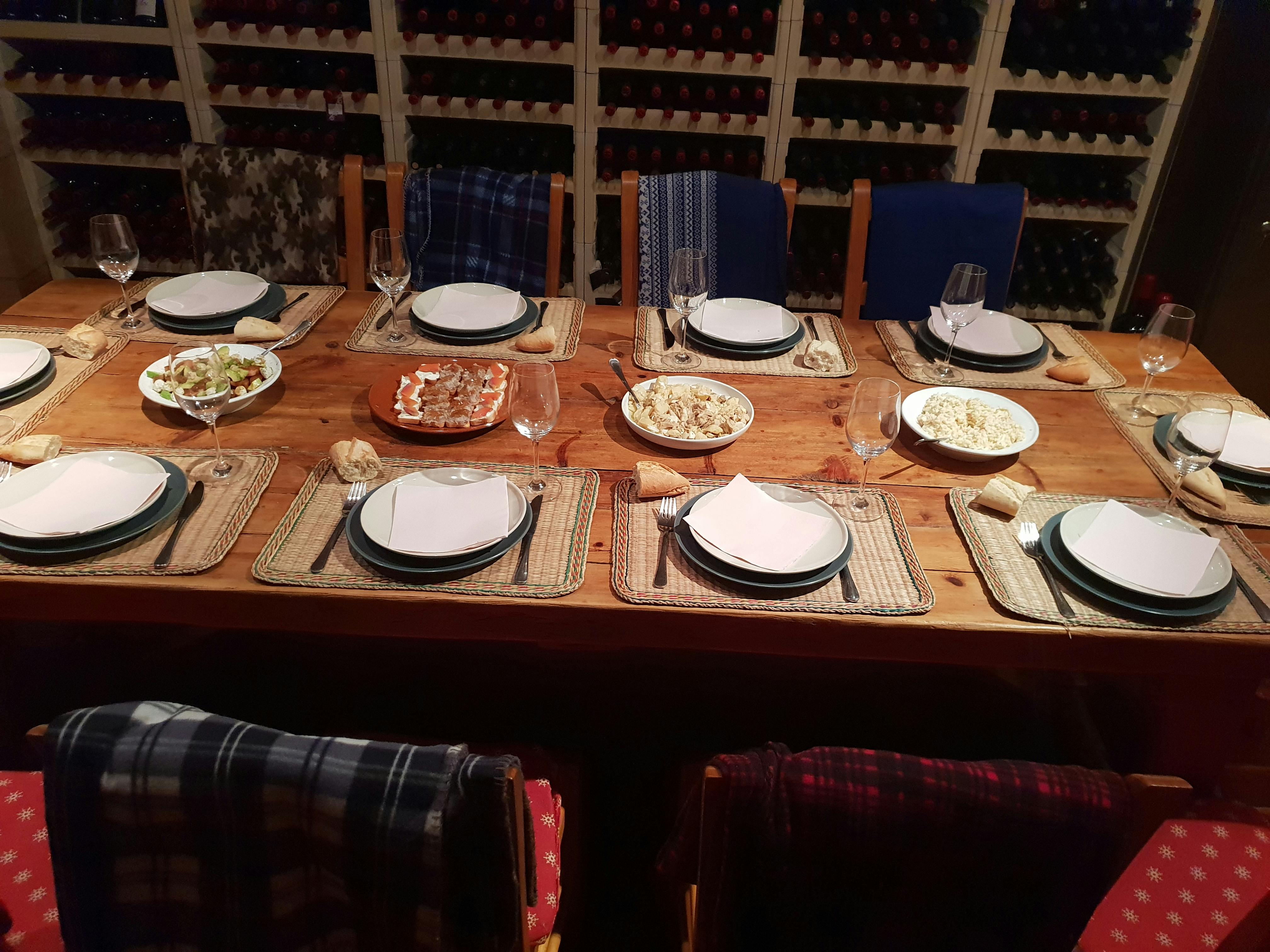 Wooden dining table set for six with plates, cutlery, glasses, and several bowls of food. Wine racks are seen in the background.