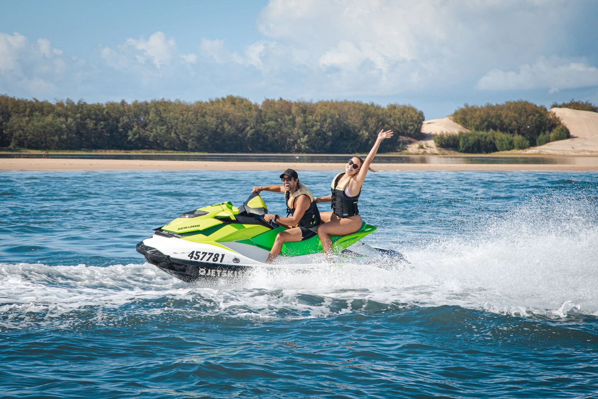 Two people riding a green jet ski in open water, one raising an arm. A distant sandy shore with trees is visible in the background.