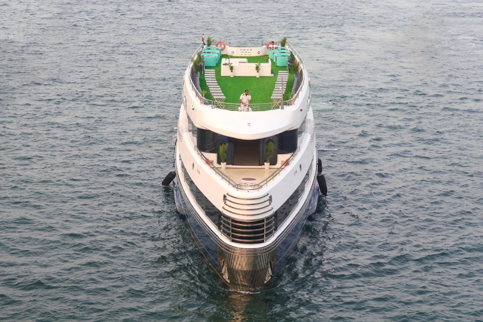 Front view of a luxury yacht with multiple decks, green turf, and a couple standing on the upper deck, surrounded by water.