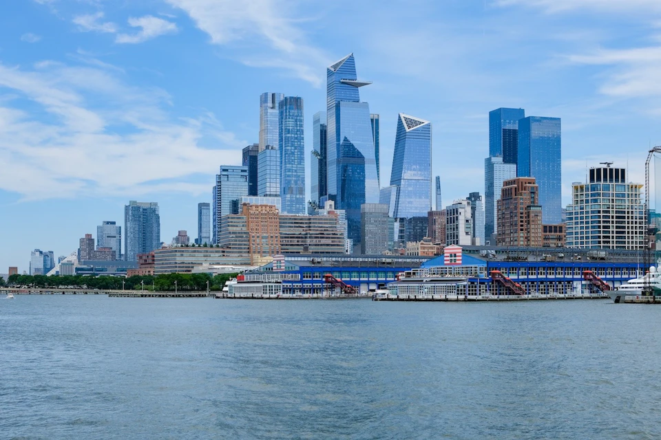 City skyline with modern skyscrapers and waterfront buildings, viewed from across a body of water under a blue sky.