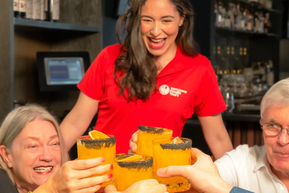 A group of people raising orange-colored drinks in a toast, with a smiling server in a red shirt in the background.