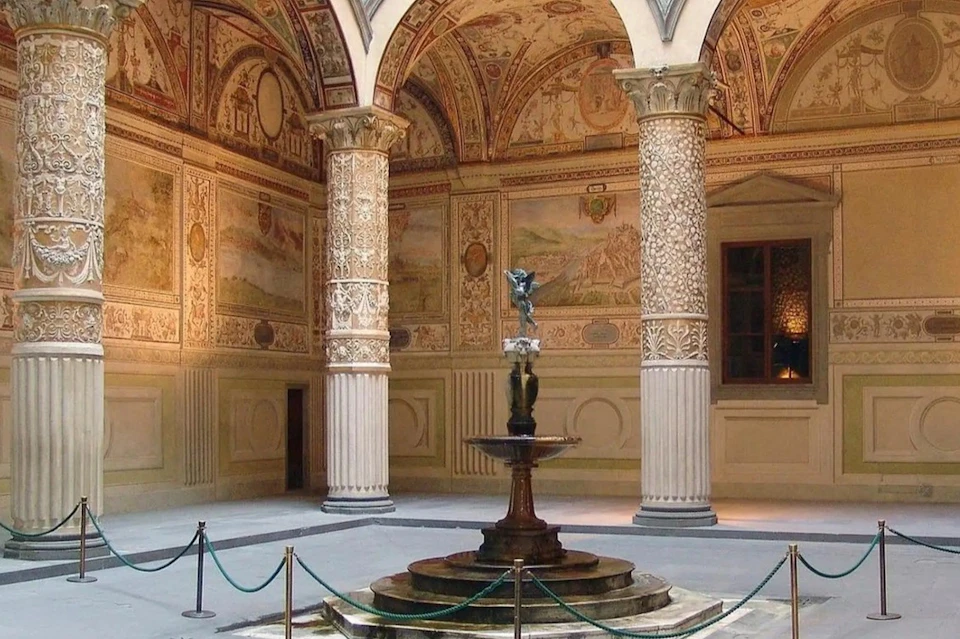 A historic courtyard with ornate columns, frescoes on walls, and a central fountain, enclosed by roped barriers.