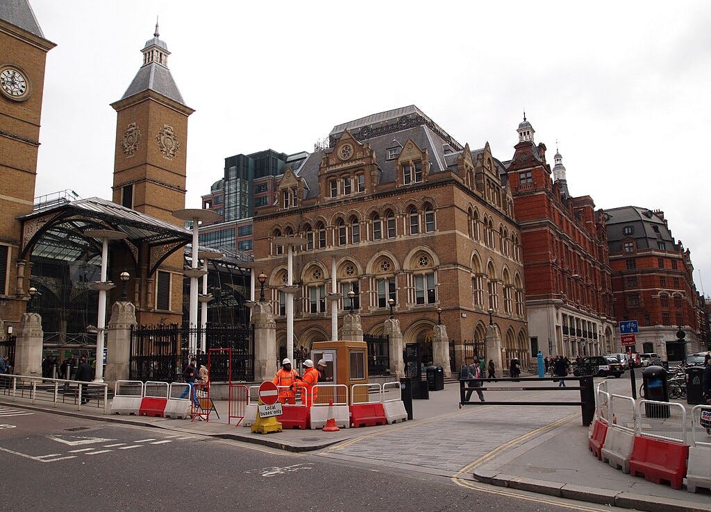 Straßenszene mit historischen Gebäuden, Uhrentürmen, Spaziergängern und Bauarbeitern in der Nähe von Schranken und einem Schild "Straße gesperrt".