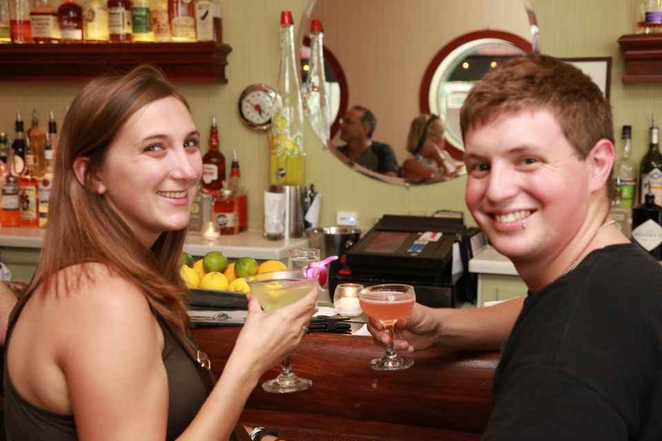 A smiling person at a bar holding a cocktail, with various bottles, a mirror, and another person visible in the background.