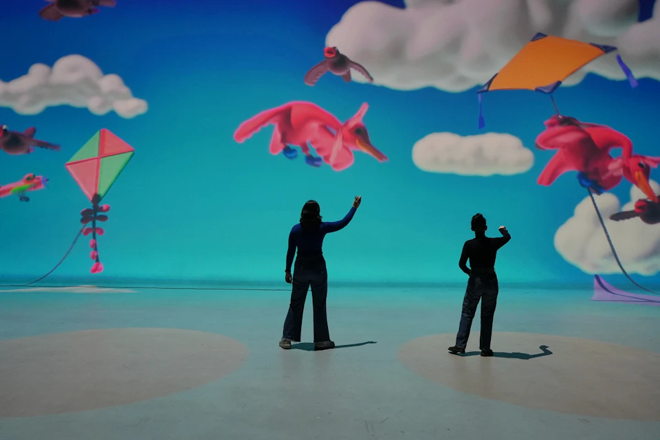 Two people interact with a digital display featuring colorful animals, clouds, and shapes against a blue background.
