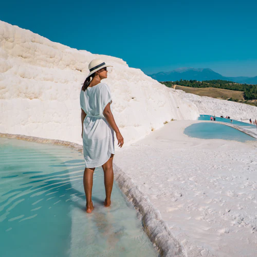 En kvinna i vit klänning och hatt går i grunt turkost vatten omgiven av vita terrasserade formationer, med människor i bakgrunden.