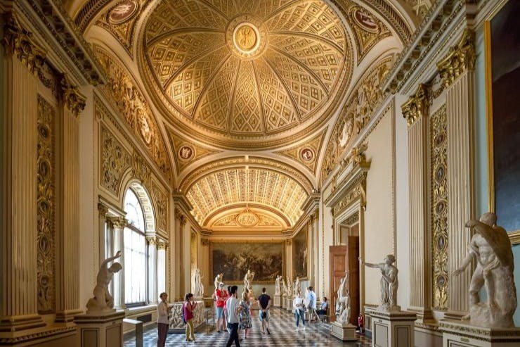 정교한 아치형 천장과 커다란 아치형 창문이 있는 화려한 홀에서 관광객들은 고전적인 조각품을 감상합니다.