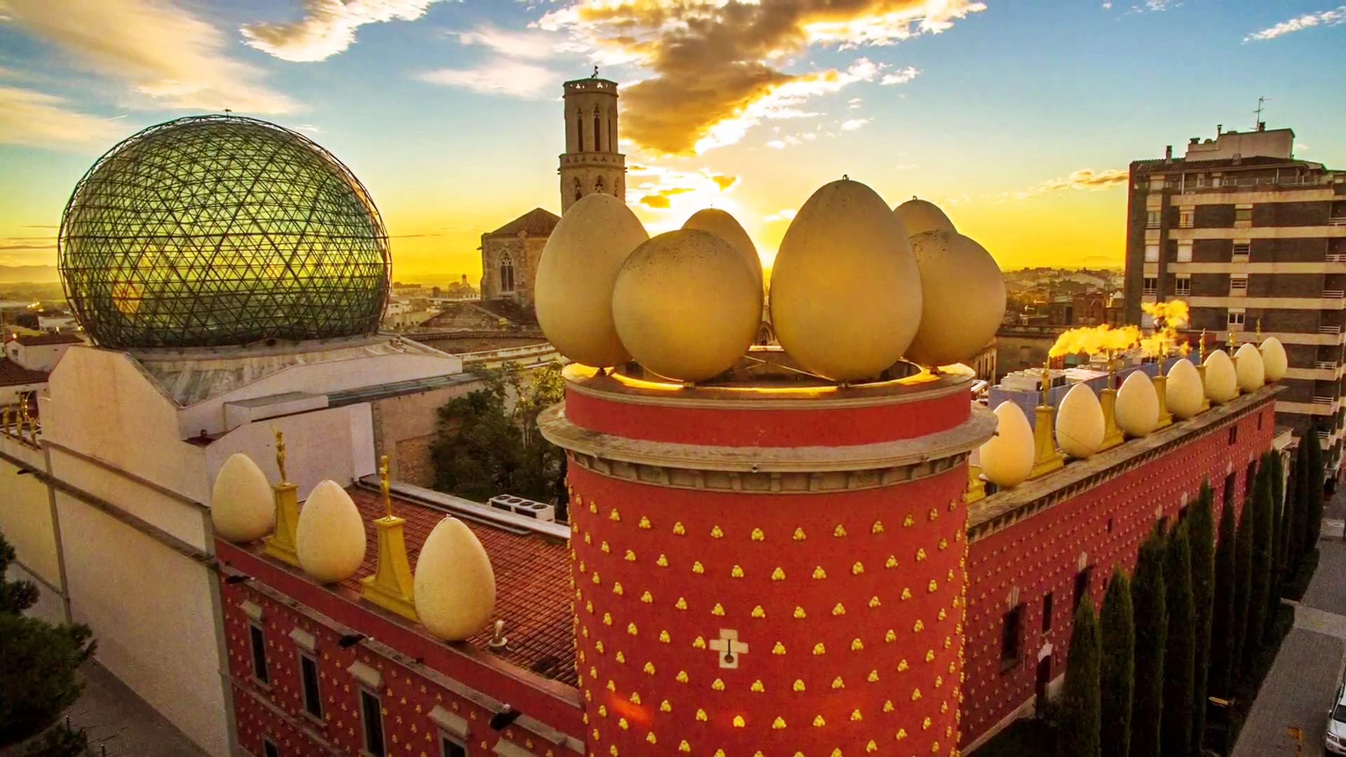 Red building with large egg sculptures on the roof, a glass dome, and a church tower against a sunset sky in the background.