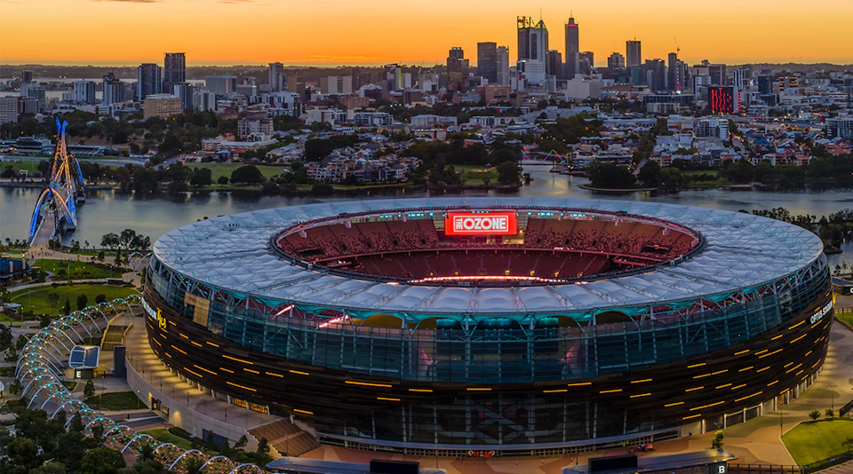 Aerial view of a stadium with a digital screen reading "THE OZONE," city skyline and sunset in the background.