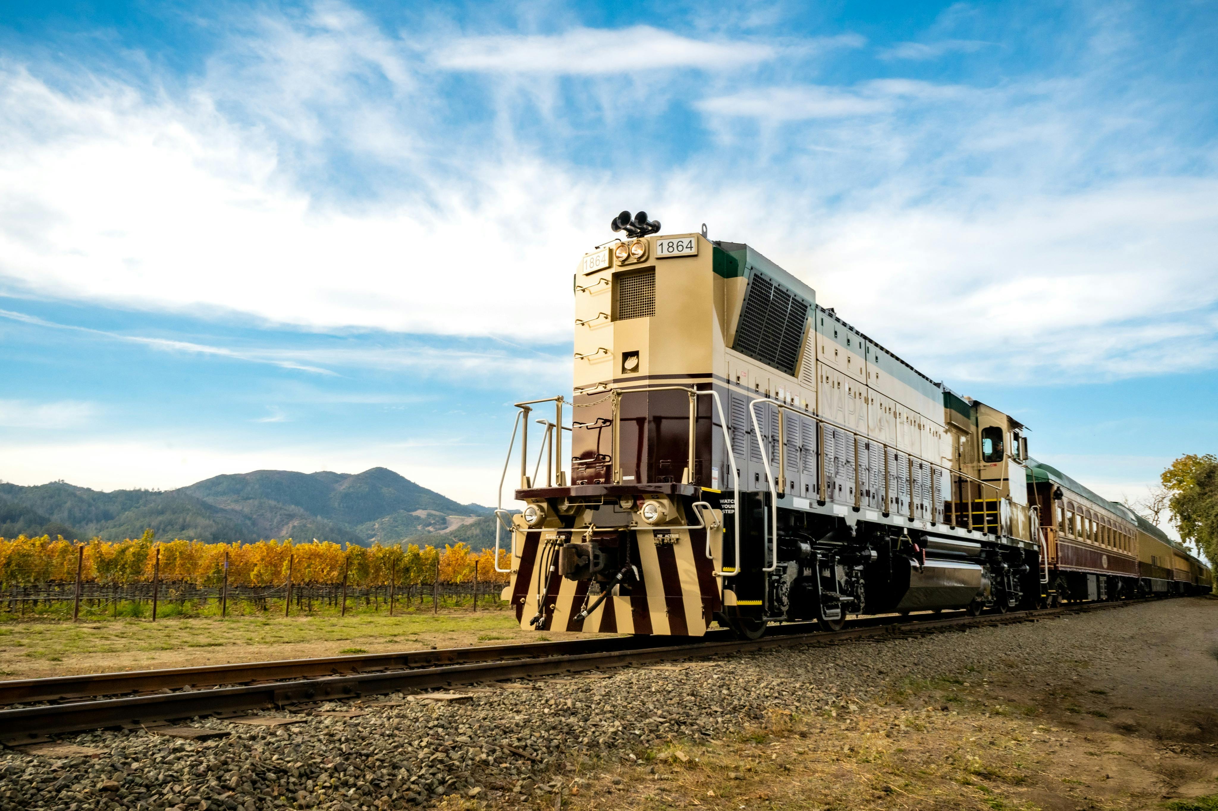 A beige locomotive traveling on tracks by a vineyard, with mountains and a blue sky in the background.