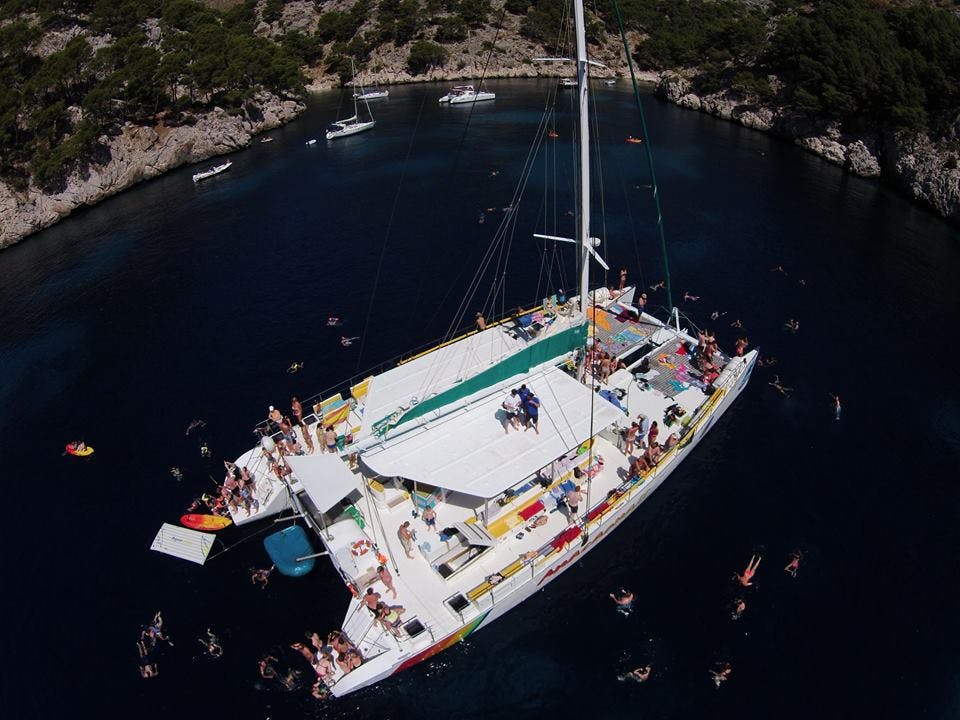 Un grand catamaran dans une baie avec de nombreuses personnes à bord et d'autres nageant dans l'eau bleue environnante. Voiliers en arrière-plan.