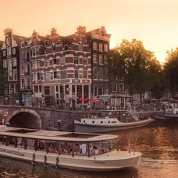 En kanalbåt navigerar en vattenväg framför klassiska Amsterdam-byggnader och träd under en gyllene solnedgång.