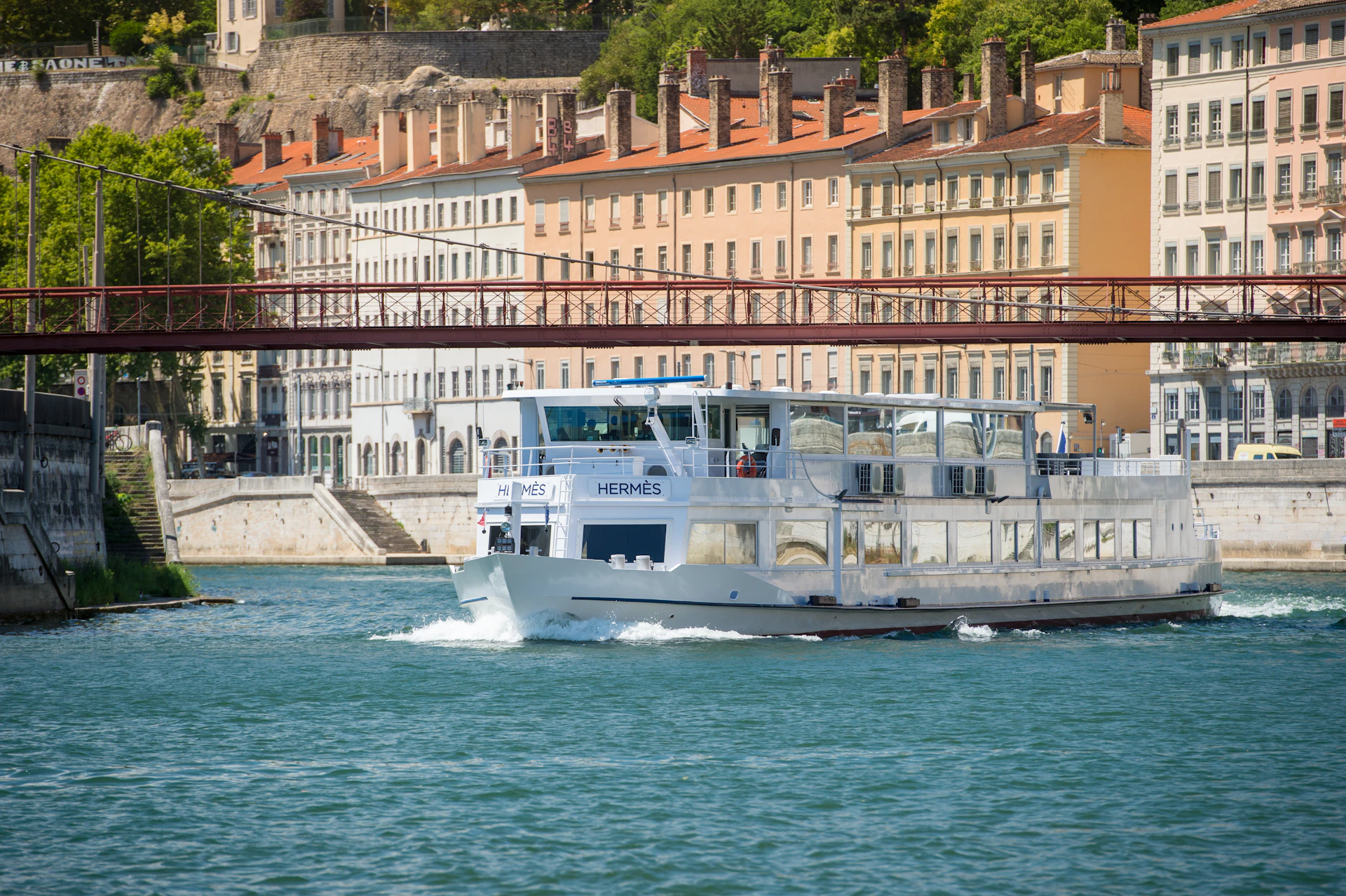 A white riverboat named "Hermès" travels on a river, with colorful historic buildings and a red pedestrian bridge in the background.