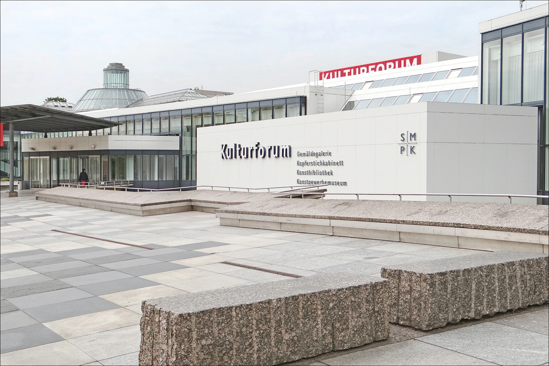 Modern building complex with large "Kulturforum" sign, gray concrete exterior, and expansive paved plaza in front.