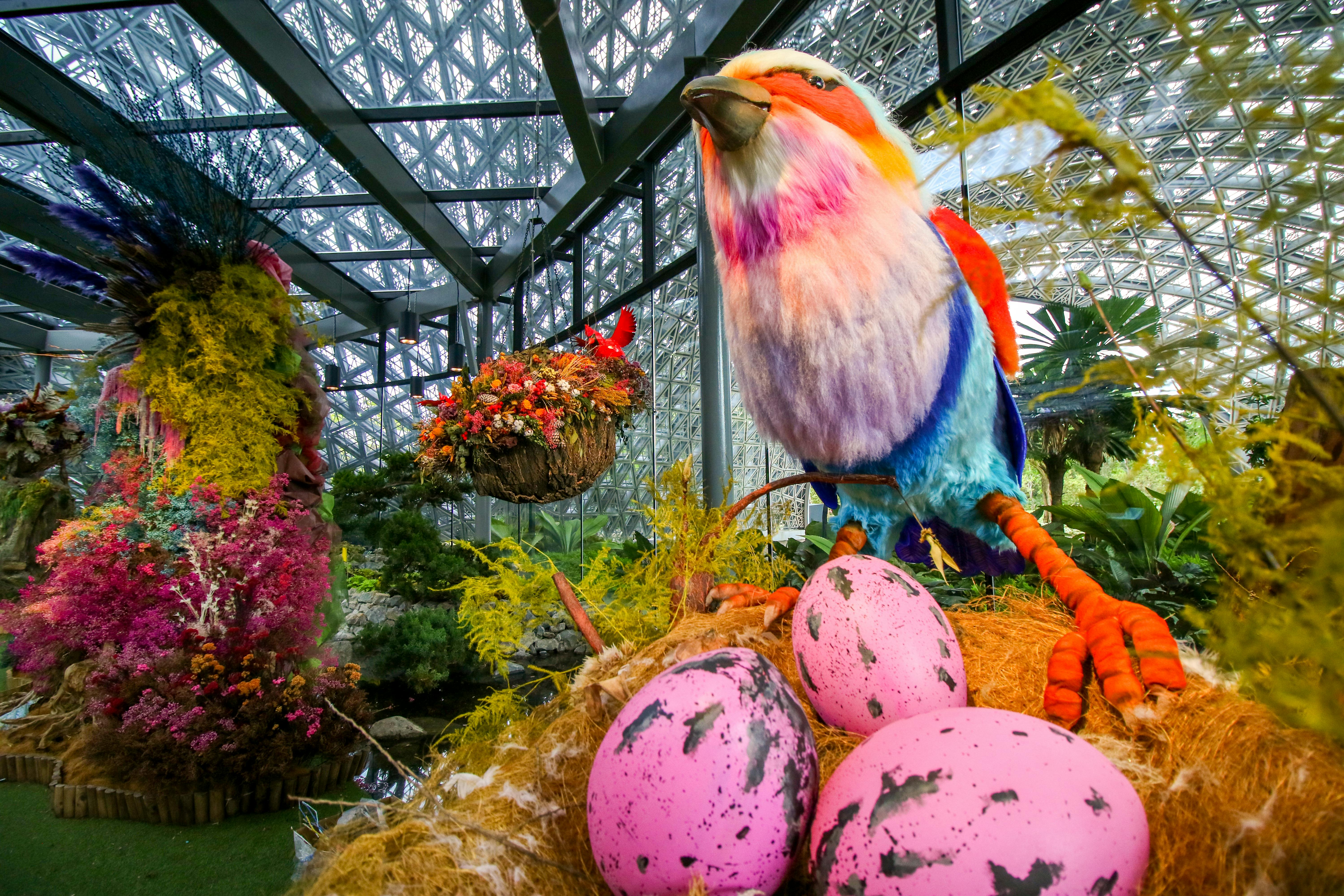 Large colorful bird statue with pink speckled eggs in a nest, surrounded by plants inside a glass-roofed conservatory.