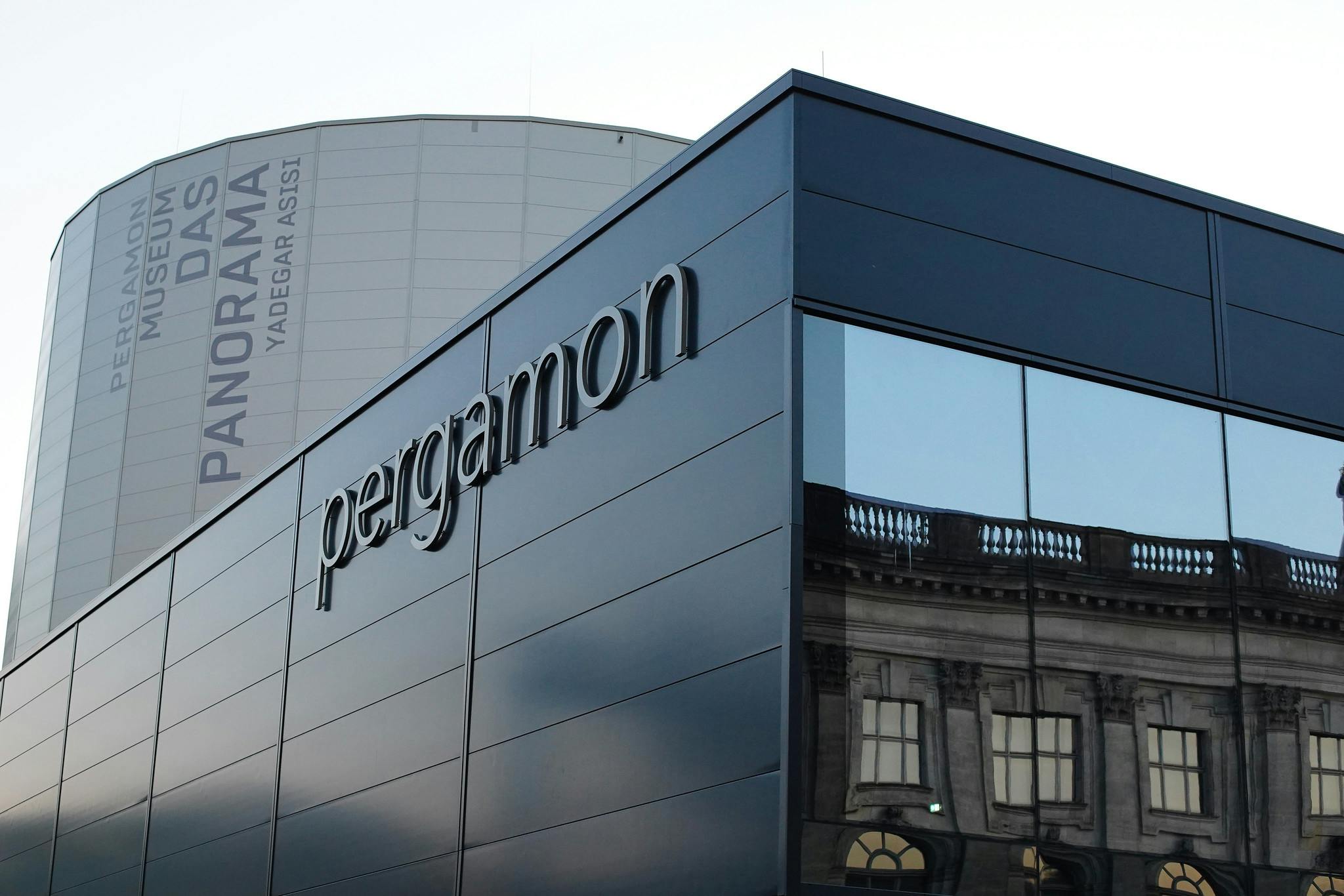 &#34;페르가몬 박물관 다스 파노라마&#34; 라고 적힌 배경에 &#34;페르가몬&#34; 간판, 반사창, 원통형 구조가 있는 현대적인 건물입니다.