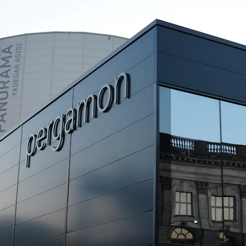 Modern byggnad med "Pergamon" -skyltar, reflekterande fönster och en cylindrisk struktur i bakgrunden märkt "Pergamon Museum DAS PANORAMA."