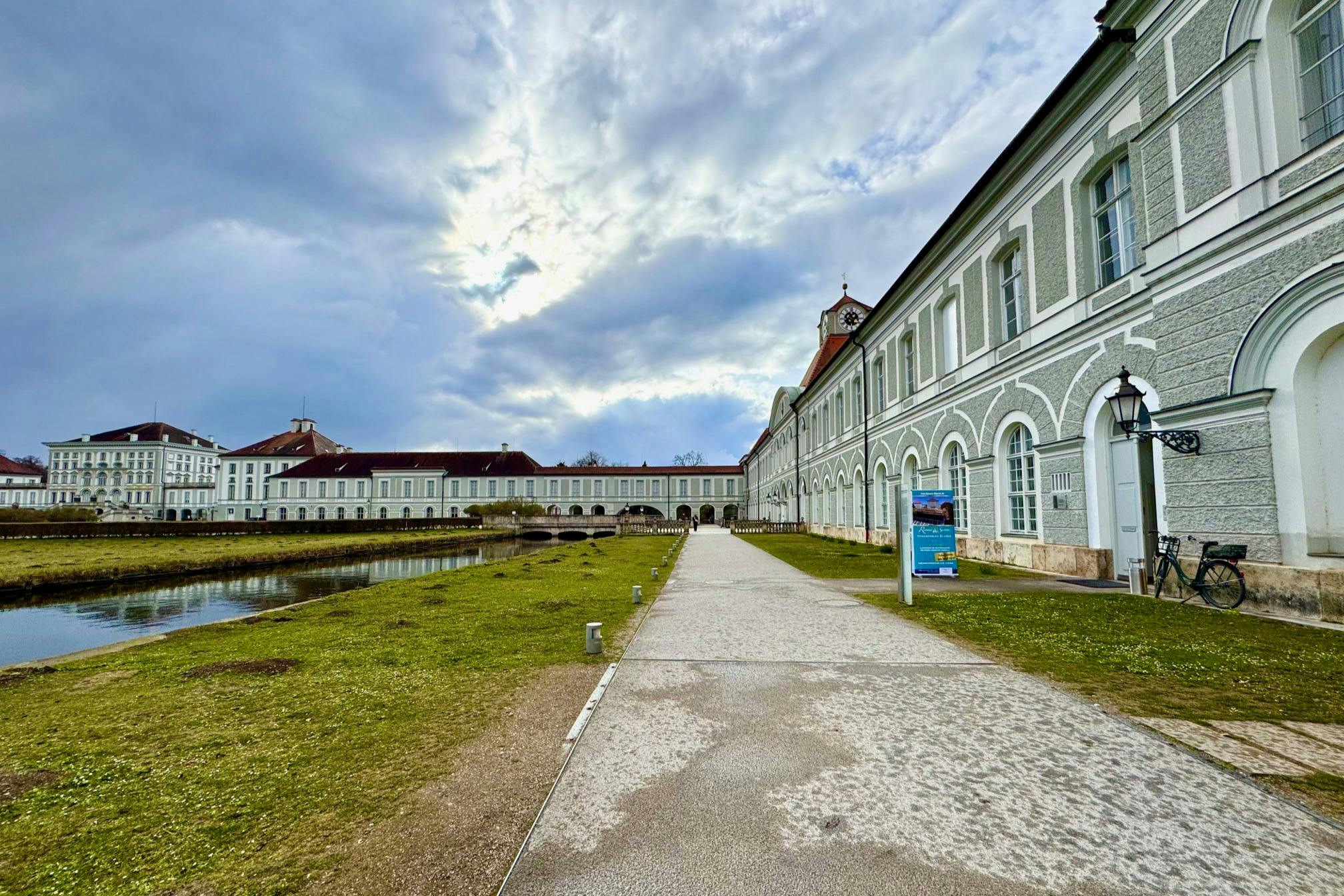 A historical building with a long pathway leading to it, surrounded by grass and water, under a partly cloudy sky.