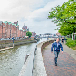 En man går längs en flodväg med tegelbyggnader och en gångbro i bakgrunden; en röd turbåt är på vattnet.