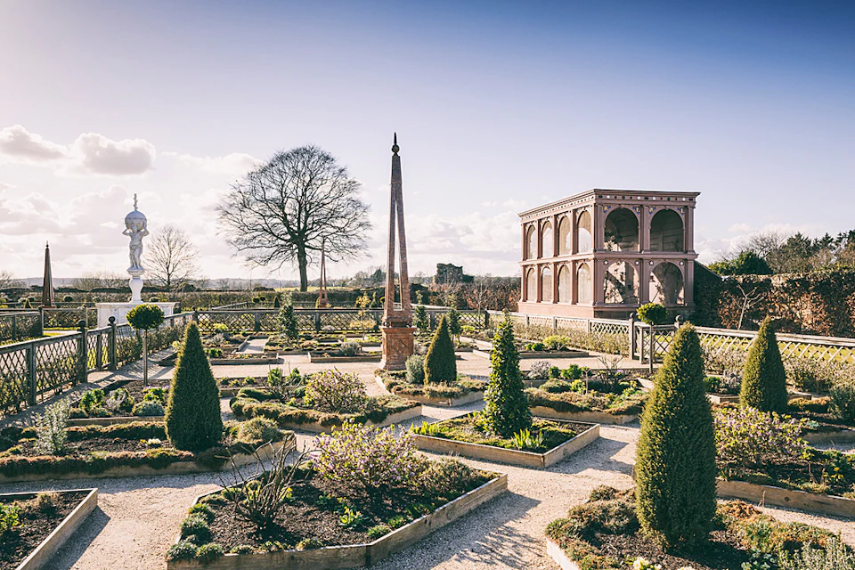 A formal garden with neatly trimmed hedges, an obelisk, and a large, open-sided stone structure under a clear sky.