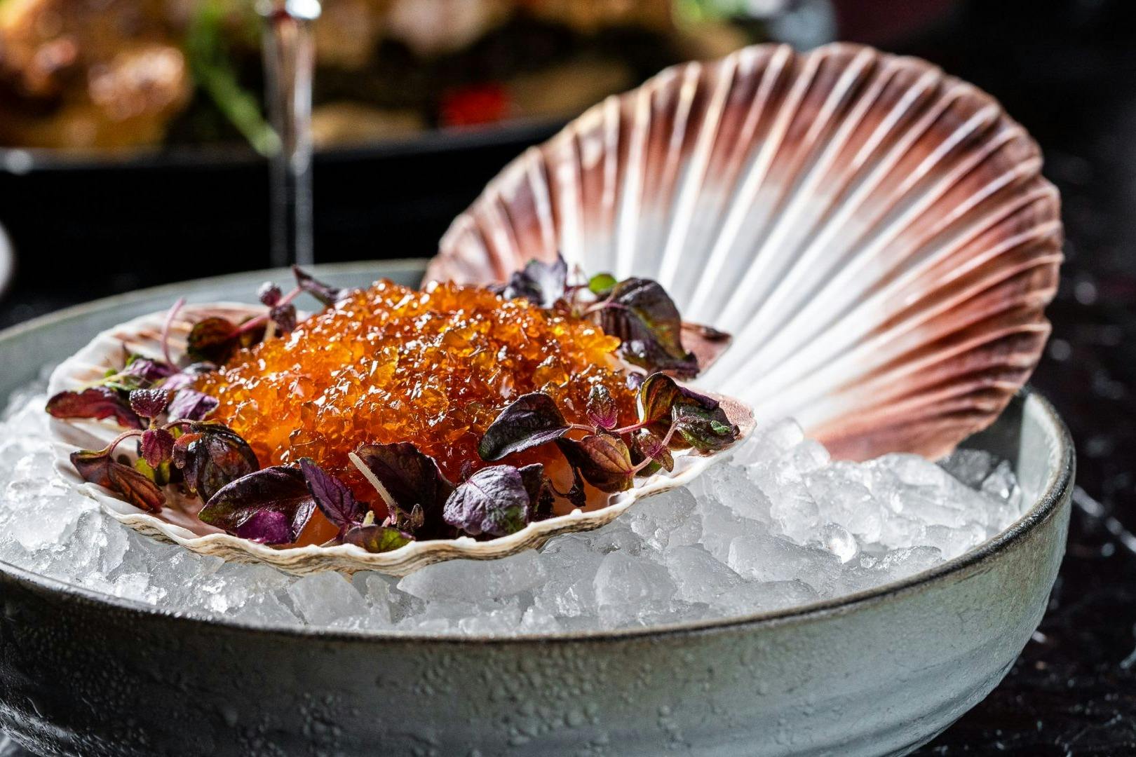 A scallop shell filled with vibrant orange roe and garnished with greens, served on a bed of crushed ice.