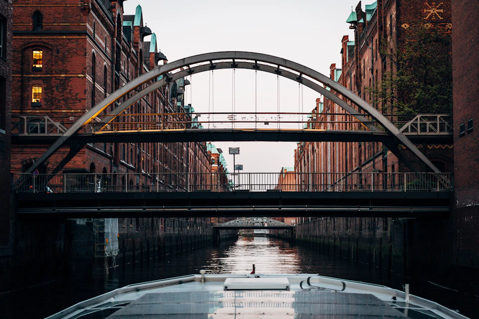 Brick buildings line a canal with multiple bridges overhead, viewed from a boat.