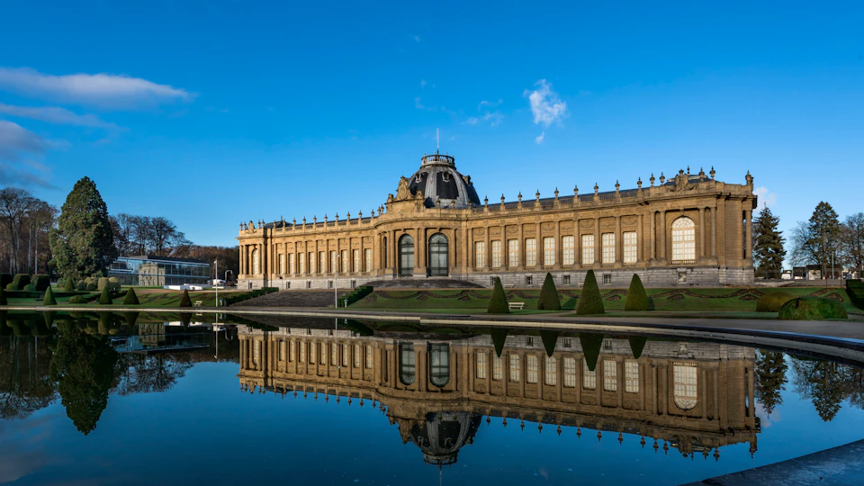 Ornate historic building with a dome, large windows, and well-manicured shrubs, reflected in a calm pond under a clear blue sky.