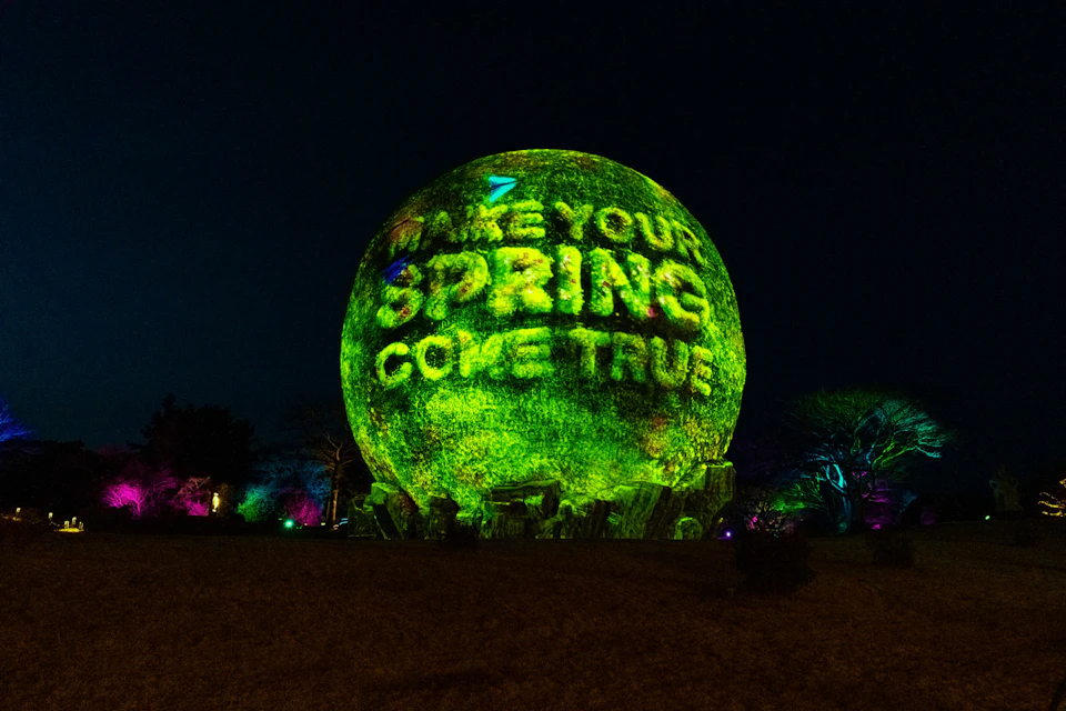 A spherical structure illuminated with green light displaying the text 'Make Your Spring Come True' at night.