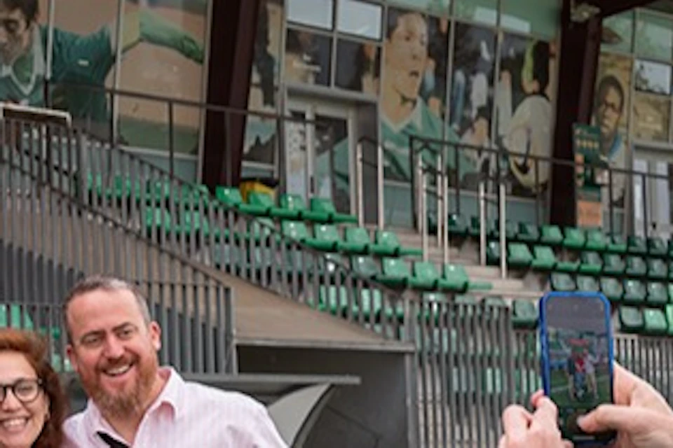 A man taking a photo of three people posing on a soccer field in front of a stadium with "UE Cornellà" signage.
