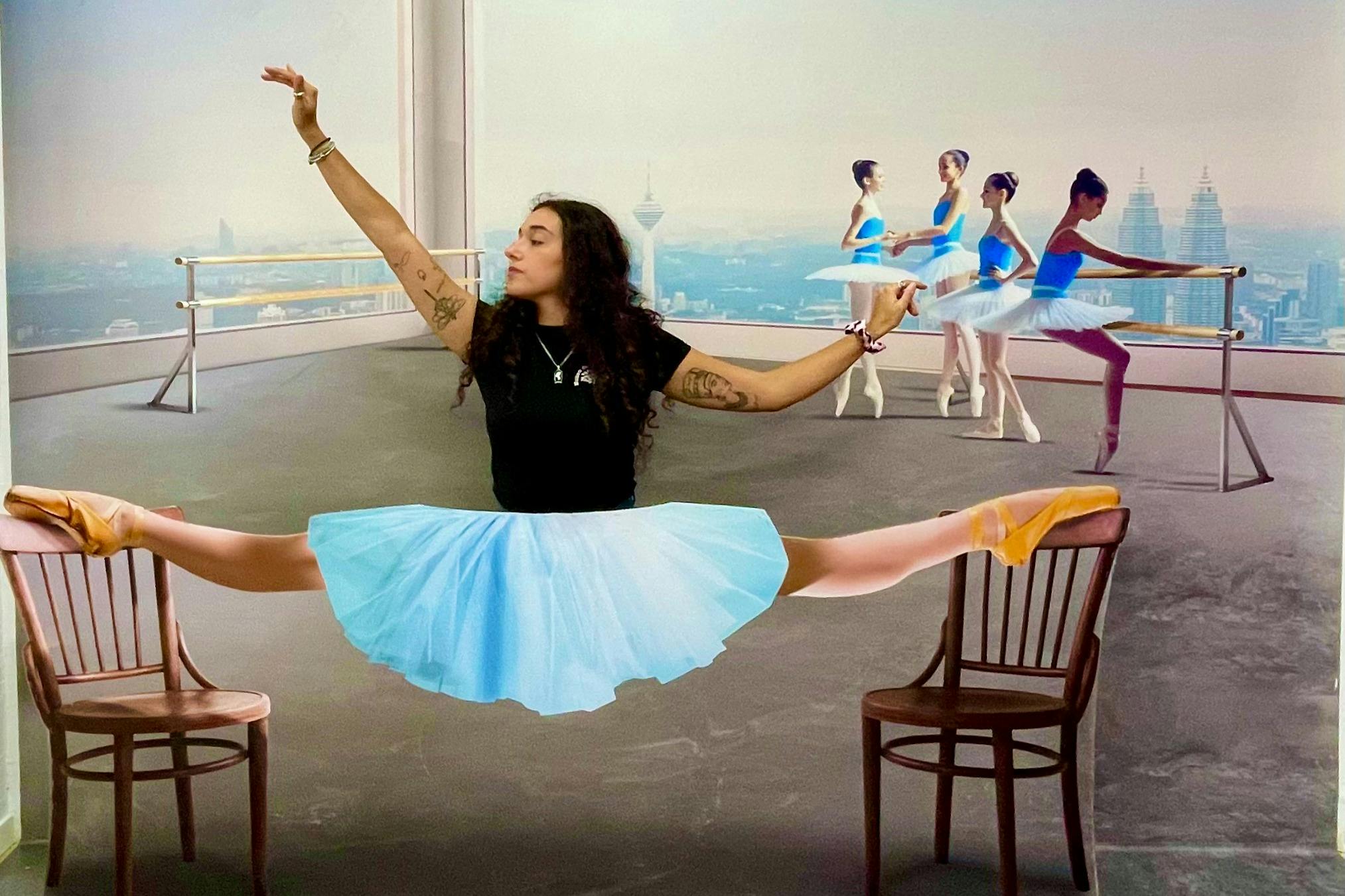 Una persona realitza una partició entre dues cadires amb un tutú blau, amb un fons d'estudi de ballet amb ballarins.