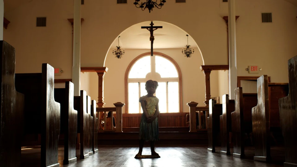 A child stands at the center aisle of a church, with pews on either side, a large cross, and arched windows in the background.