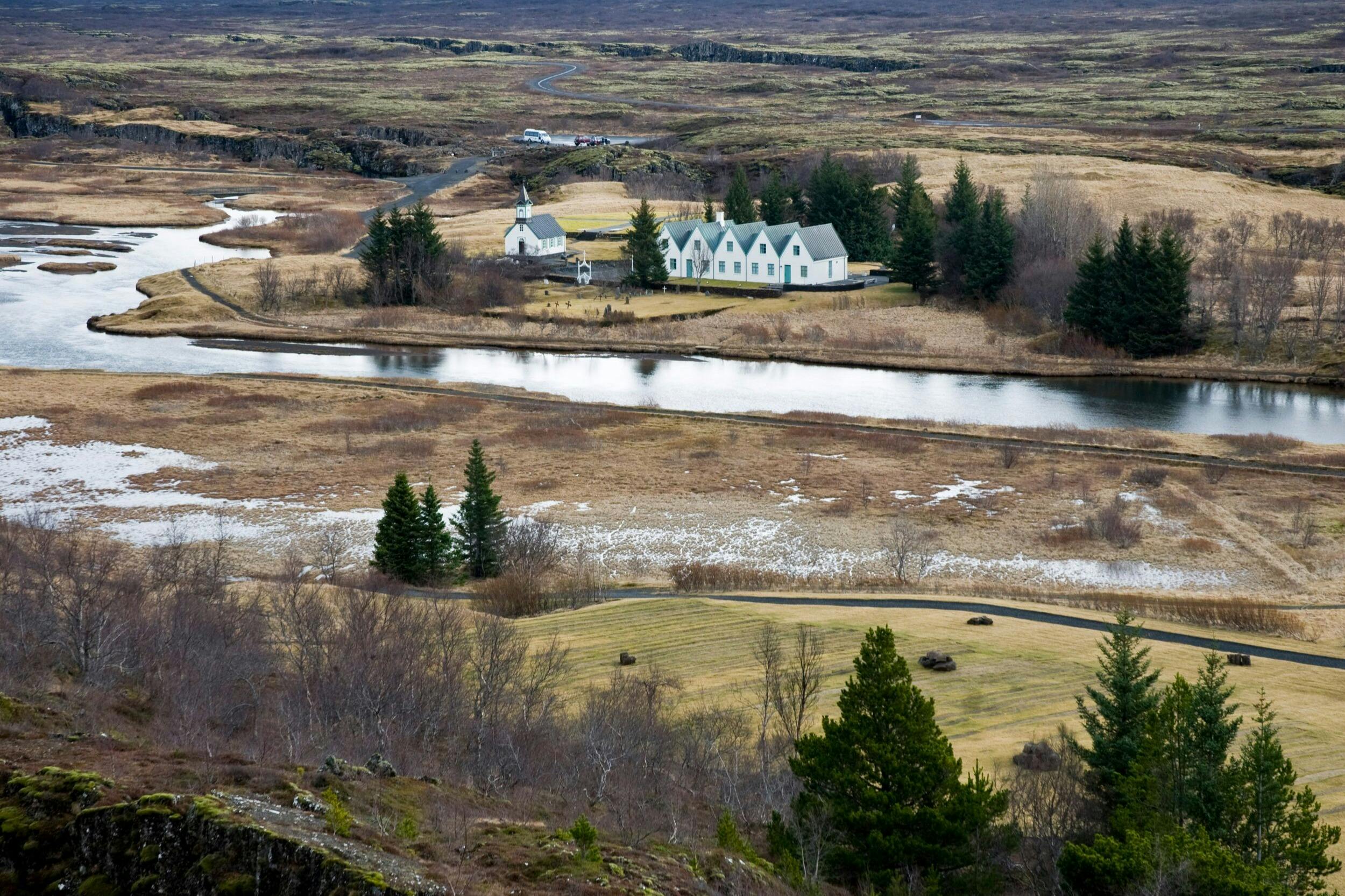 A rural landscape featuring a cluster of white buildings with green roofs surrounded by trees and a winding river.
