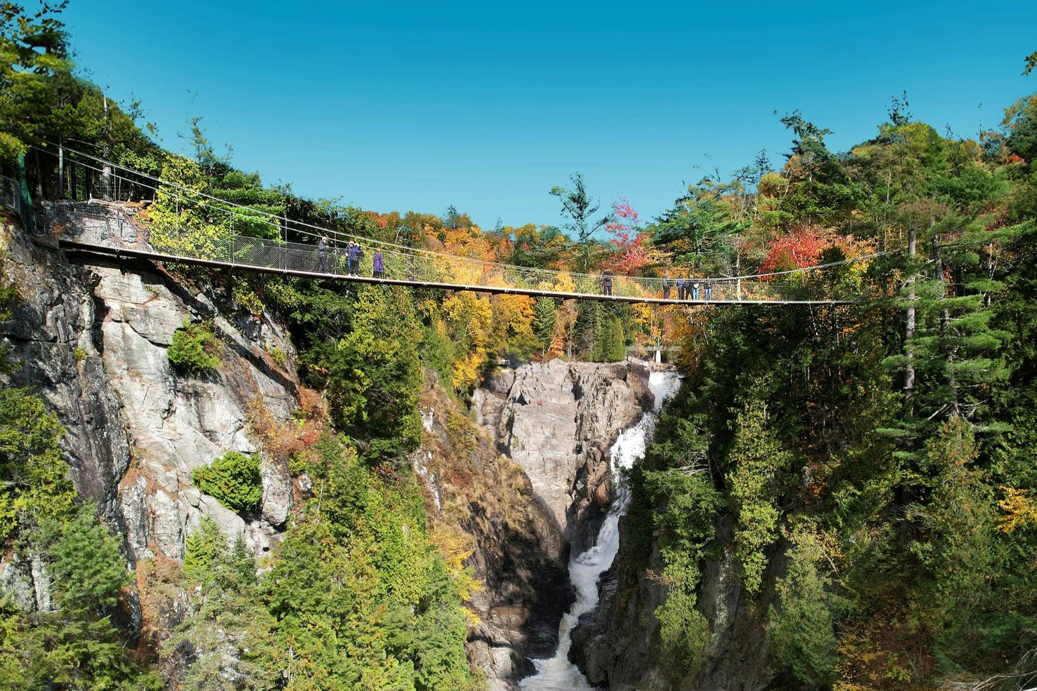 Un pont suspendu enjambe une gorge avec une chute d'eau, entourée de falaises boisées et de feuillages d'automne.
