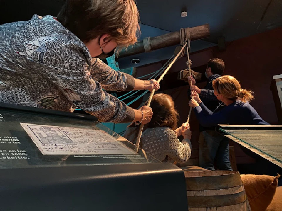Four people pull ropes attached to a large wooden mechanism in an indoor museum exhibit.