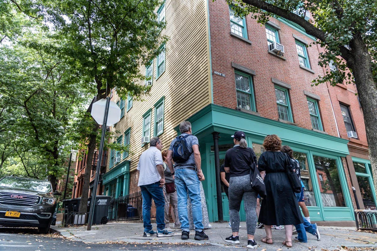 A group of people stands on a street corner, looking up at a two-story building with brick and wood siding in an urban setting.