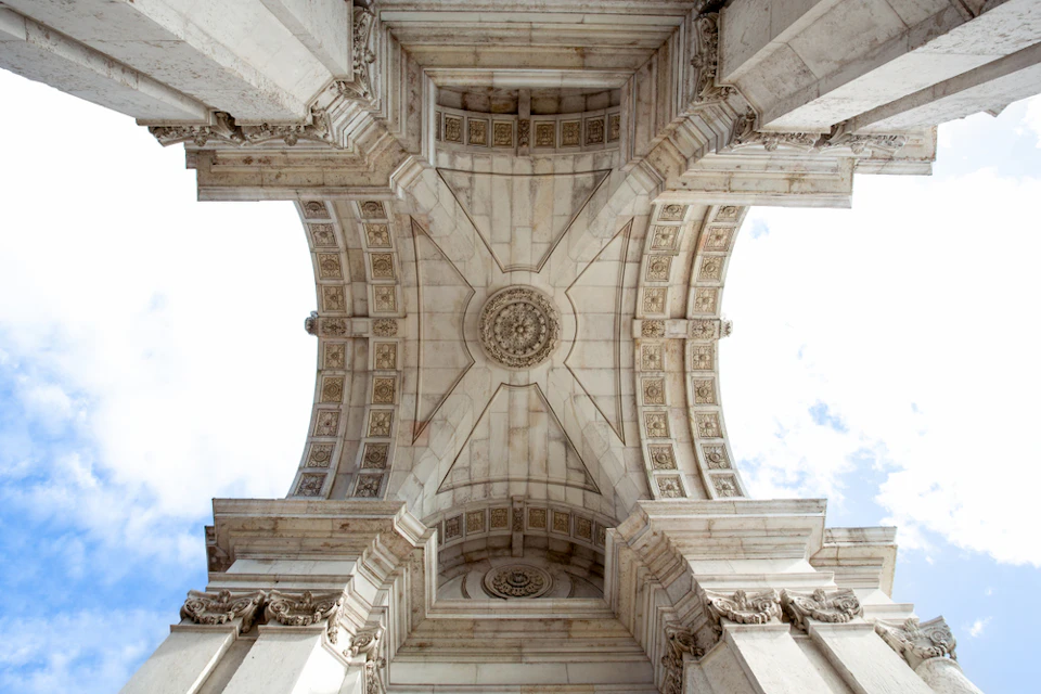 Looking up at the ornate stone ceiling of an archway with intricate carvings and architectural details against a partly cloudy sky.