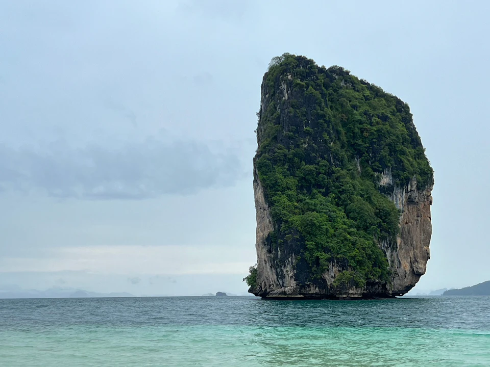 Tall, rocky island covered in greenery, standing in clear blue-green ocean under a cloudy sky.