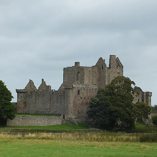 Ett gammalt stenslott med delvis förstörda murar, omgivet av träd och grönt gräs, under en molnig himmel.