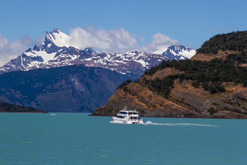 A boat cruising on a turquoise lake, surrounded by mountains with snowy peaks and hills covered in vegetation.
