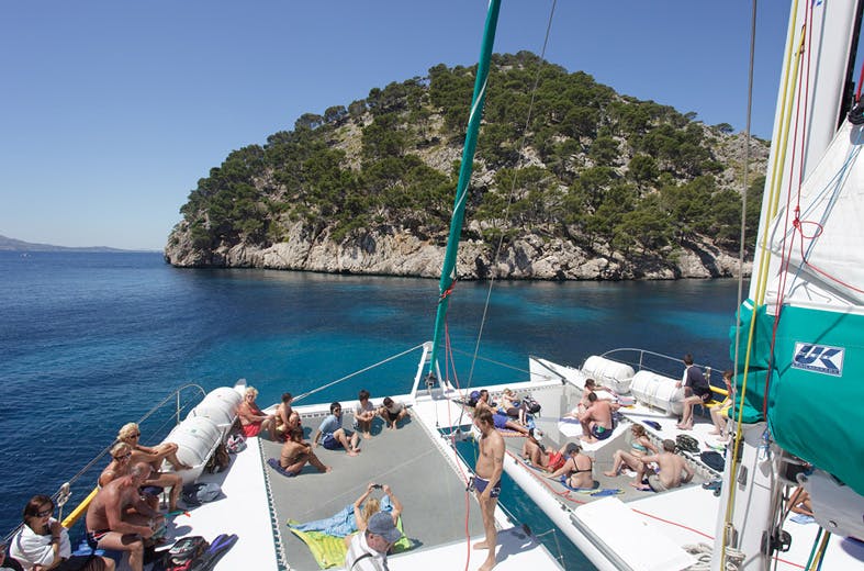 Personnes se relaxant sur un catamaran en mer, avec une île rocheuse couverte d'arbres en arrière-plan, sous un ciel bleu clair.