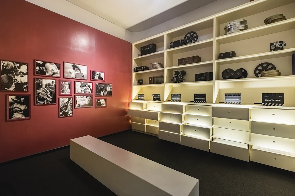 A room with shelves holding film equipment and photos on one wall, a bench in the center, and warm lighting.