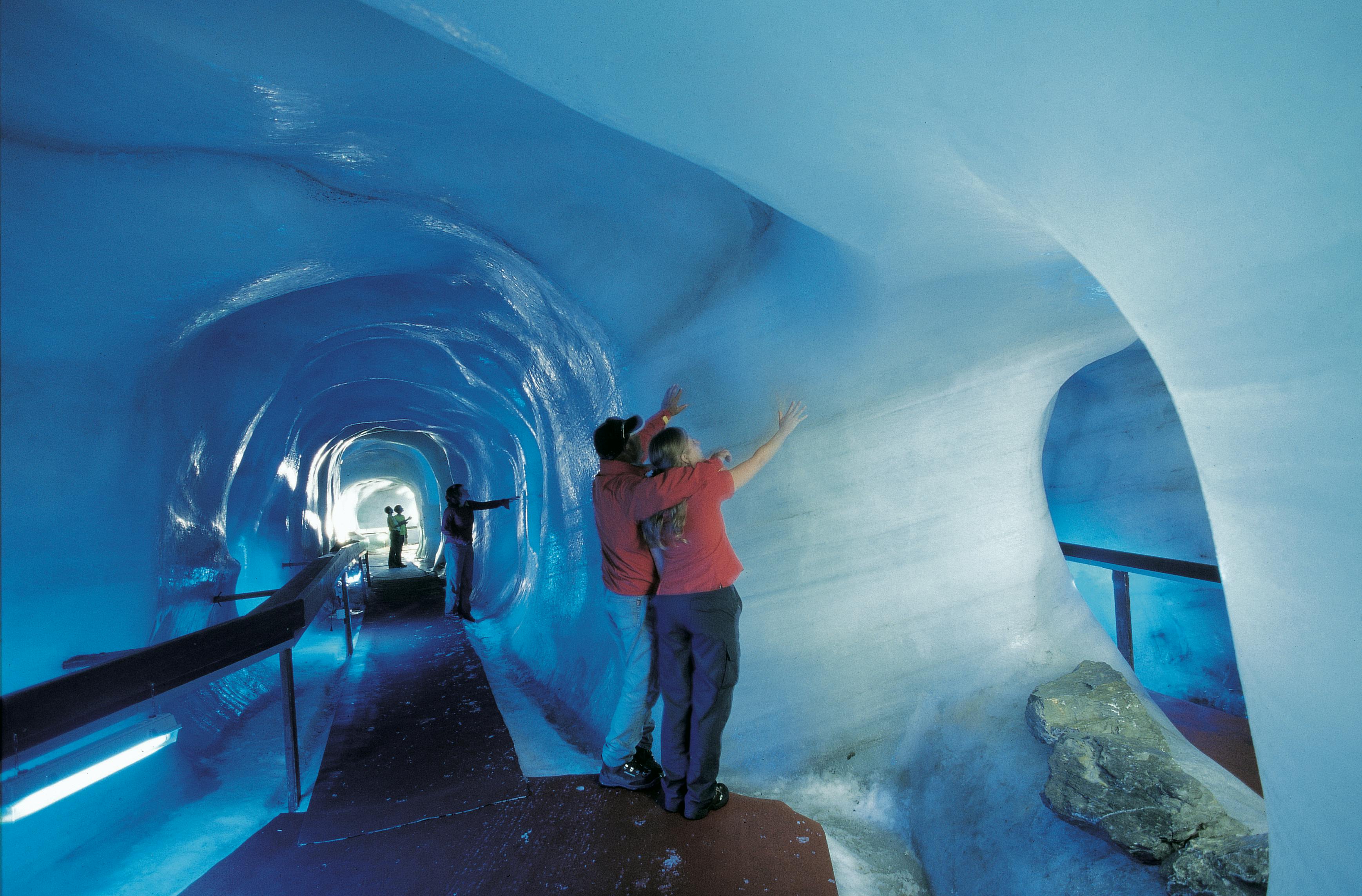 Dva lidé v červených bundách se dotýkají ledových stěn uvnitř modře osvětleného ledového tunelu a v pozadí je vidět další osoba.