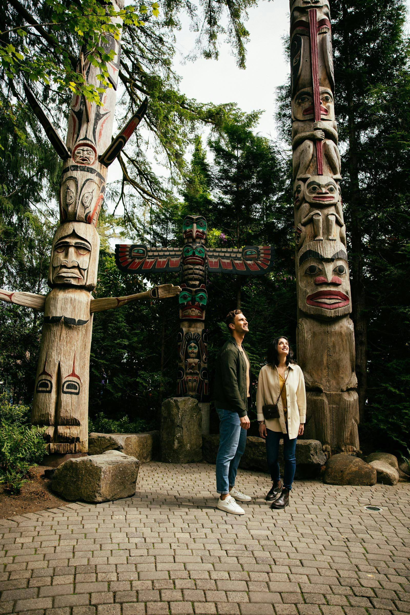 一对男女站起来，抬头看着被树木和铺好的石头包围的高大、精心雕刻的图腾柱。