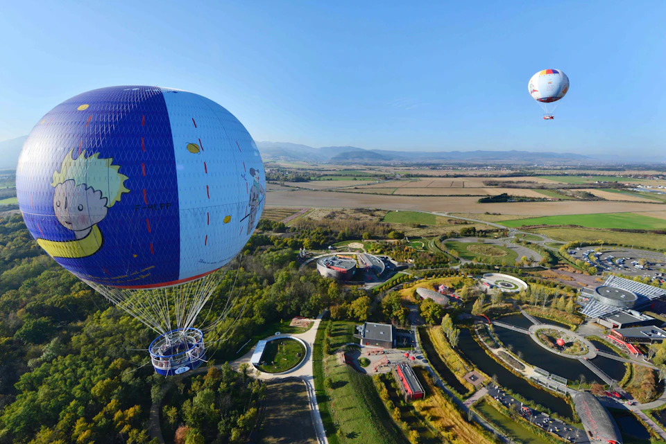 Two hot air balloons float above a park with gardens, waterways, and buildings, surrounded by a vast landscape.